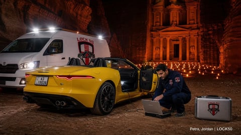 Mechanic crouching beside yellow sports car at night with white van and illuminated historic building in background